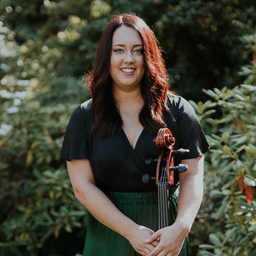 Woman standing outdoors holding a cello, smiling.