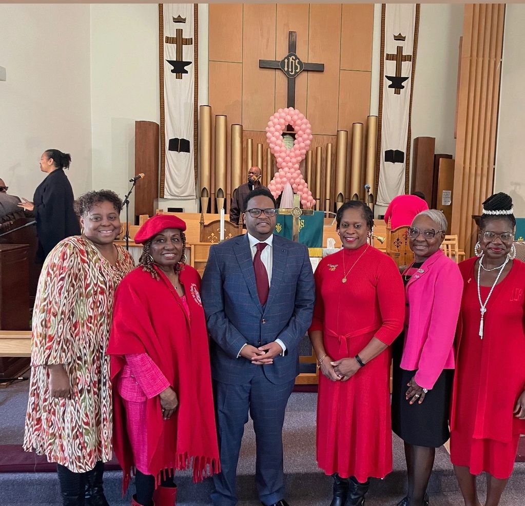 Group of six people posing in a church with pink ribbon decoration behind.