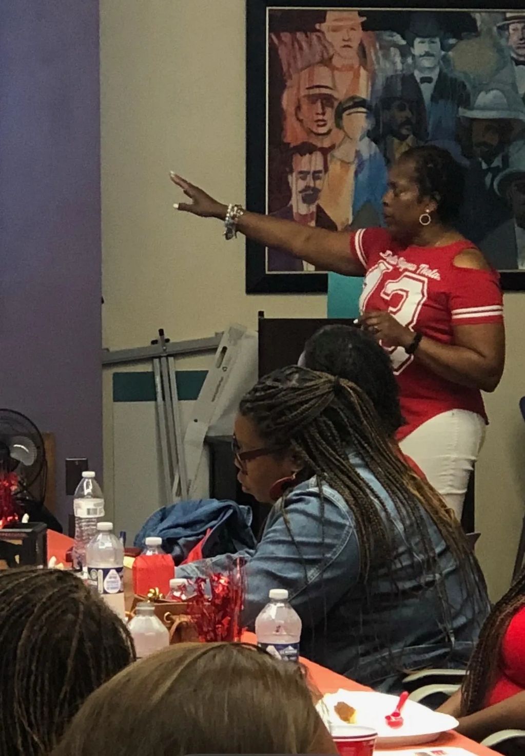 A woman in a red shirt gestures while others sit at a table with drinks and decorations.
