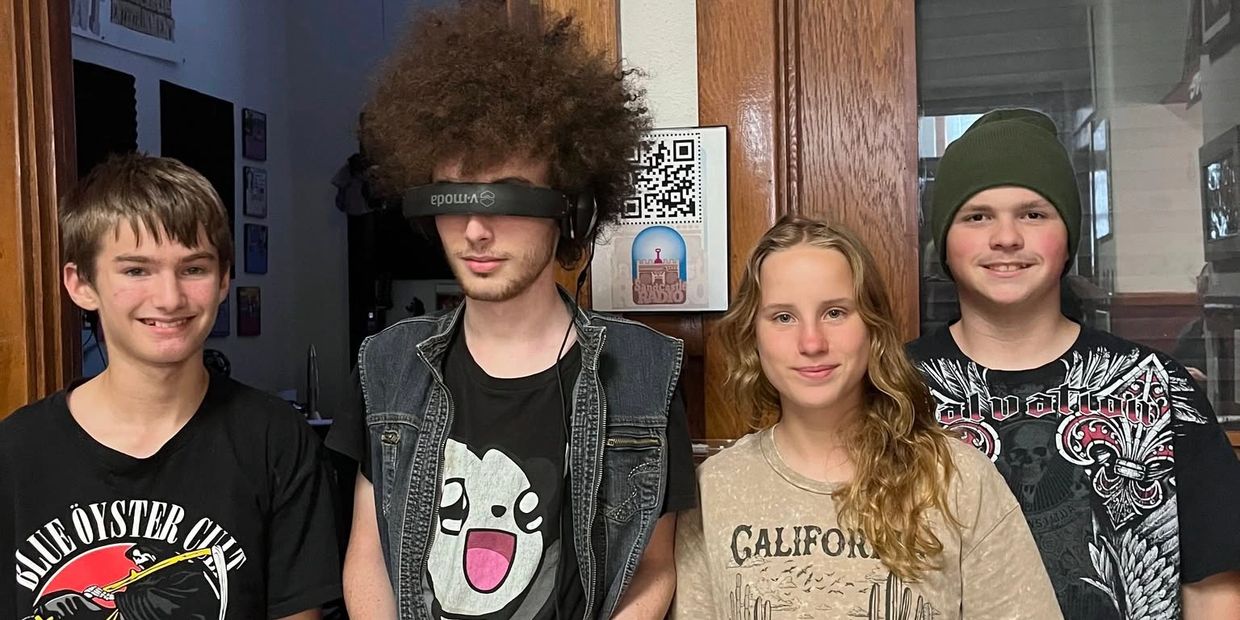 Four teenagers posing indoors, one with a VR headset and distinctive curly hair.