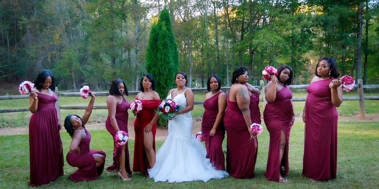 Bride in white surrounded by bridesmaids in burgundy dresses holding pink bouquets outdoors.