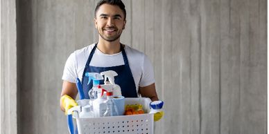 Smiling man holding a basket of cleaning supplies wearing gloves and apron.