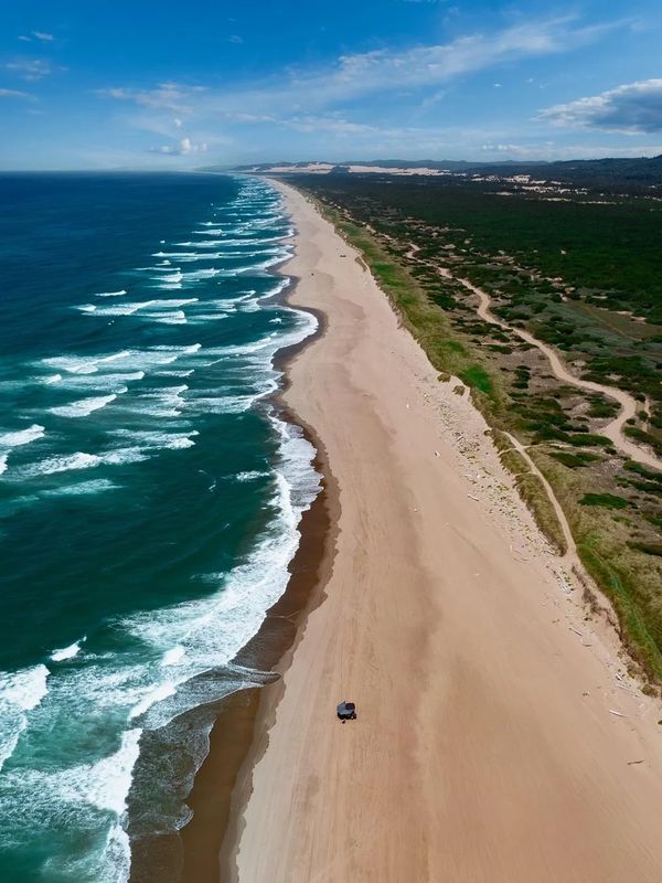 A vast beach with waves crashing and a lone vehicle on the sand.