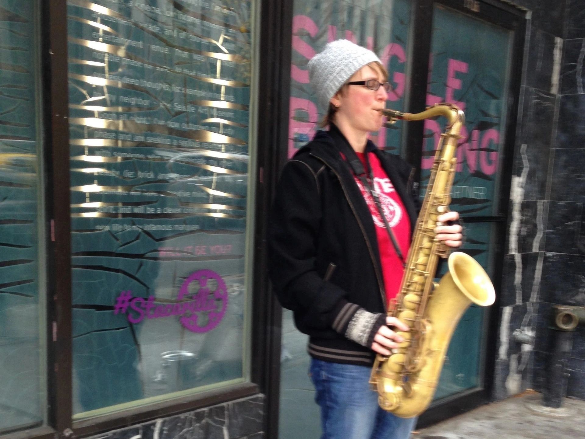 Person playing saxophone on a city street wearing a beanie and glasses.