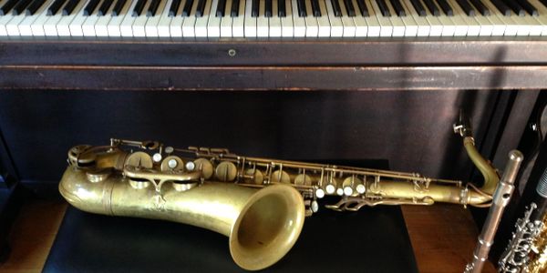 A saxophone and a flute resting on a piano bench and an old piano.