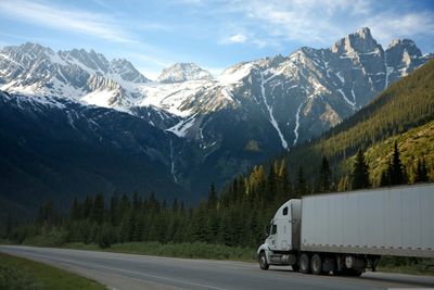 A semi-truck drives on a road beside snow-capped mountains and pine trees.