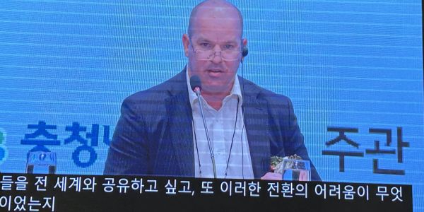 A man in a suit speaking at a conference with Korean subtitles.
