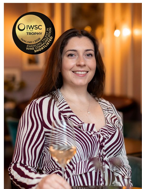 Smiling woman with striped blouse holding a glass of wine, IWSC award badge visible.