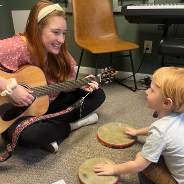 music therapist smiling and holding guitar during music therapy with young boy playing drums