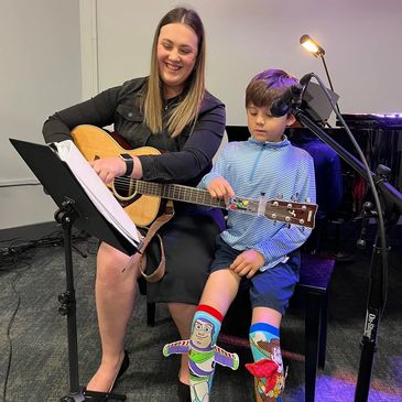 guitar teacher smiling and holding guitar while young boy presses buttons on a chord buddy.