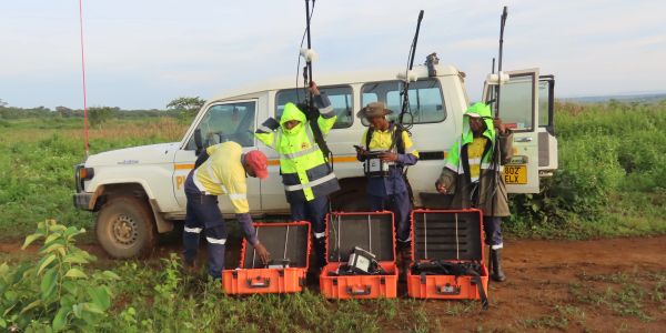 Field workers in safety gear preparing equipment near a white vehicle.