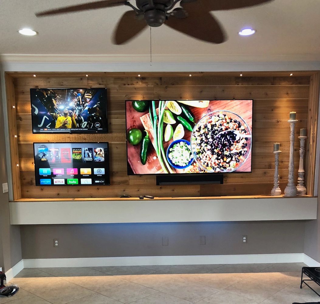 Three TVs mounted on a wood-paneled wall with decorative candles and ceiling fan.