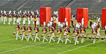 Marching band performing on a football field with red curtains in the background.