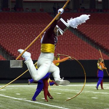 Marching band member leaps while spinning a flag on a football field.
