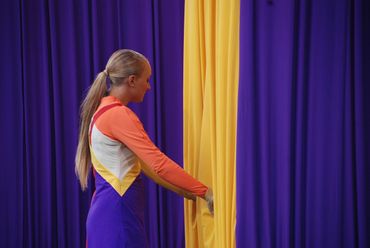 A girl in a colorful outfit pulling back a yellow curtain against a purple backdrop.