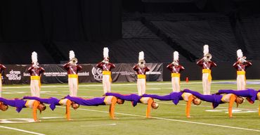 Marching band members perform a coordinated balancing act on a field.