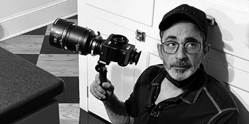 Man sitting with a camera and a large lens, wearing glasses and a cap indoors.