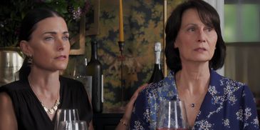 Two women sitting closely, one comforting the other at a dining table with wine glasses.