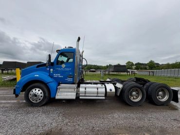 Blue semi-truck cab parked on a gravel road under cloudy sky.