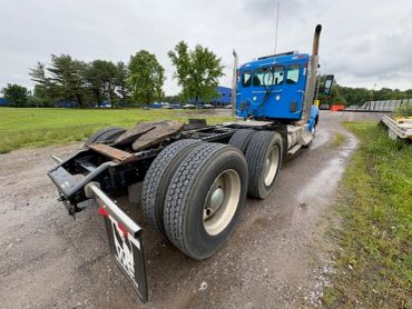 Blue semi-truck cab parked on a gravel road with a green field and trees in the background.