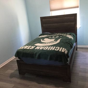Single bed with Michigan State blanket in a minimally furnished room.