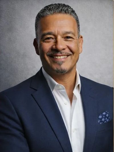Confident man in navy suit smiling against a gray background.