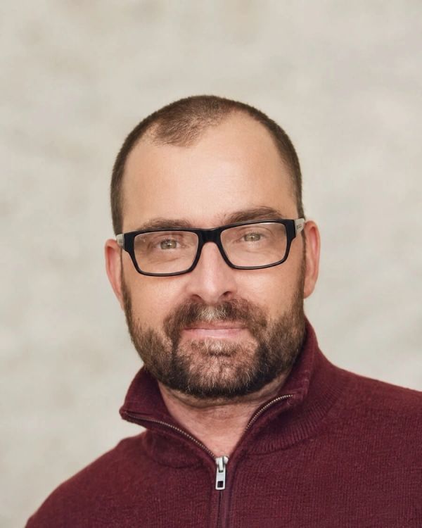Man with glasses and beard smiling with a neutral background.