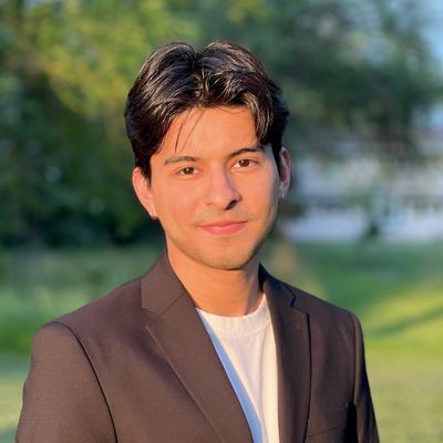 Young man in a blazer smiling outdoors with sunlight on his face.