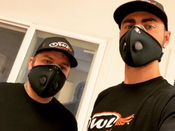 Two men wearing black masks and matching black shirts with logos.