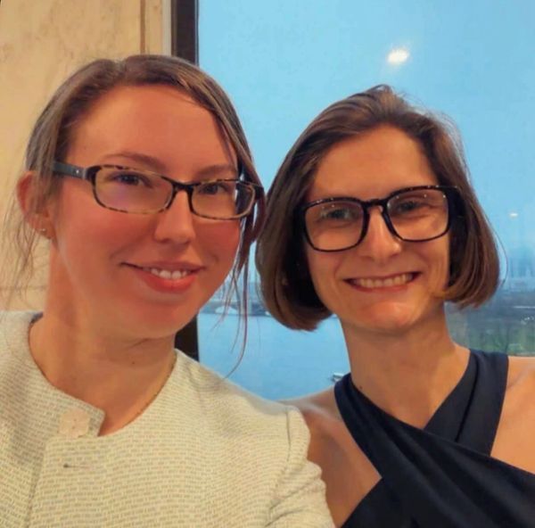 Two women wearing glasses smiling together indoors near a window.