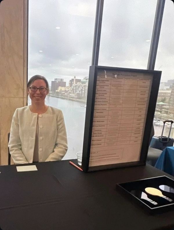 A woman in glasses and white jacket sits at a table with a large menu or sign beside her.