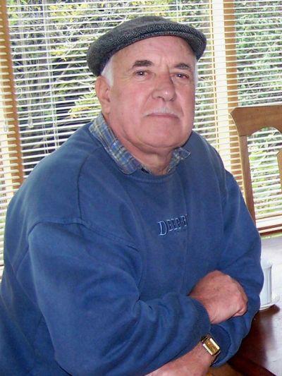 Elderly man wearing a flat cap and blue sweatshirt sitting indoors.