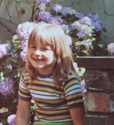 Smiling young girl in a striped shirt sitting outdoors with flowers behind her.