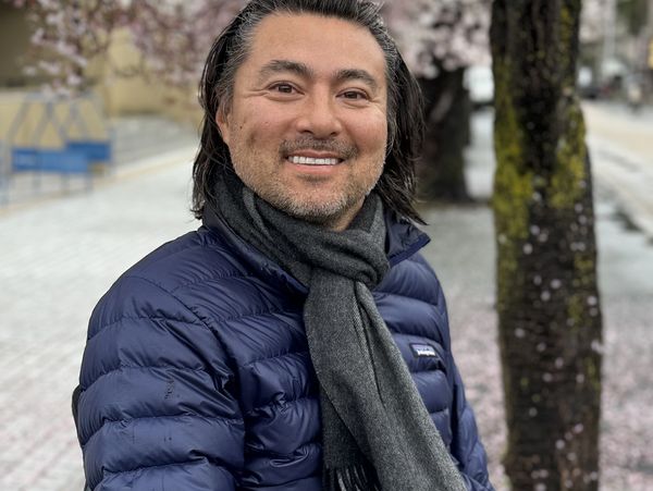 Man in blue jacket and gray scarf smiling outdoors by trees.