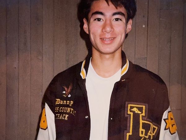 A young man smiling, wearing a letterman jacket with a wooden wall background.