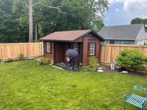 A small wooden garden shed with purple flowers in a green backyard.