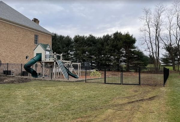 A fenced playground with slides next to a large brick house on a cloudy day.
