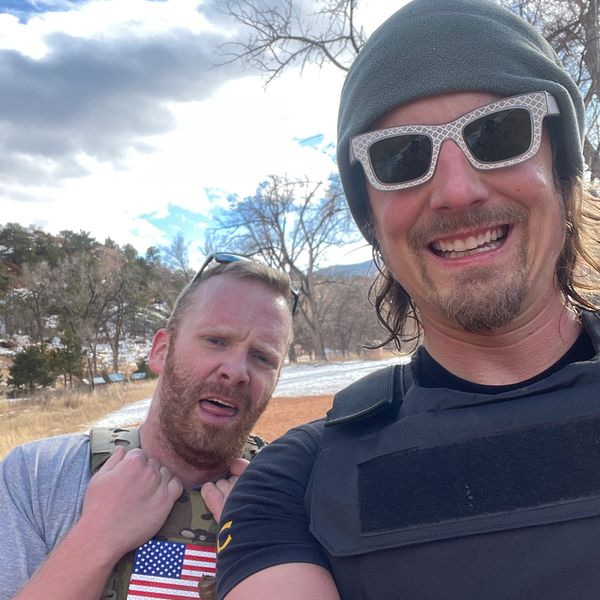Two men, one smiling and one concerned, outdoors with trees and sky.