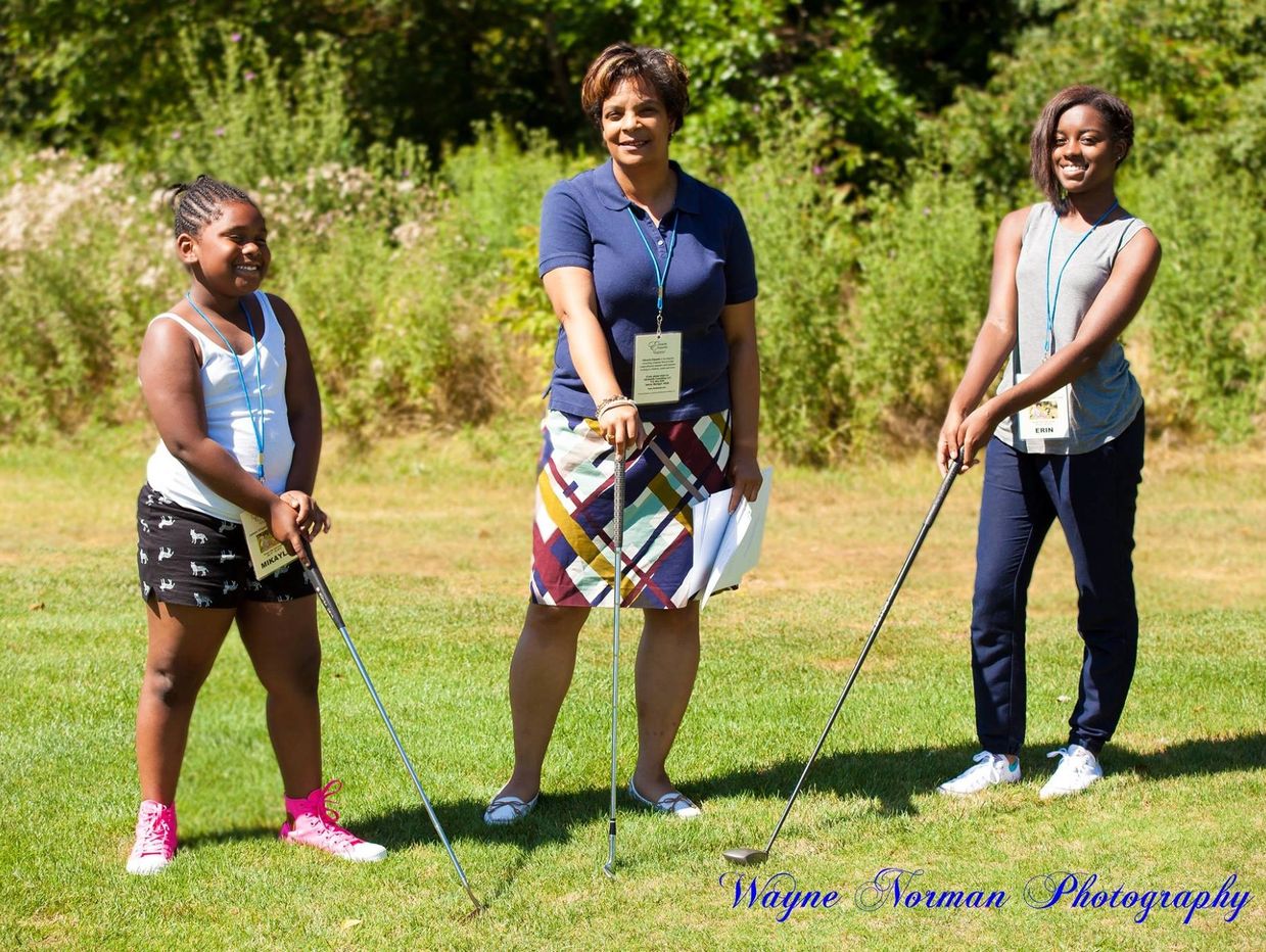 Three females smiling with golf clubs on a green lawn.