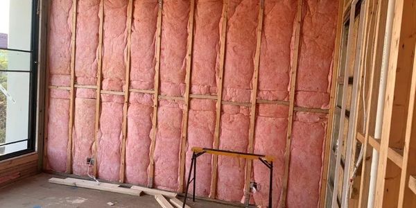 Pink fiberglass insulation installed between wooden wall studs in a construction site.