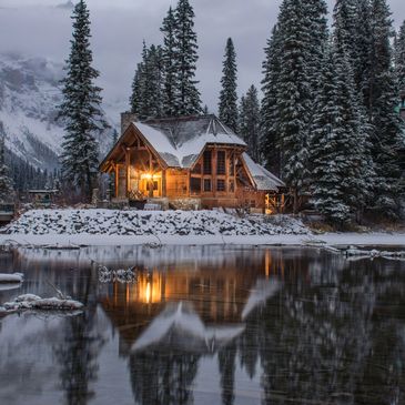 Cozy cabin with warm lights in a snowy forest reflected in a calm lake.