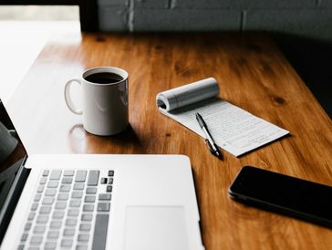 Workspace with laptop, coffee, notepad, pen, and smartphone on wooden table.