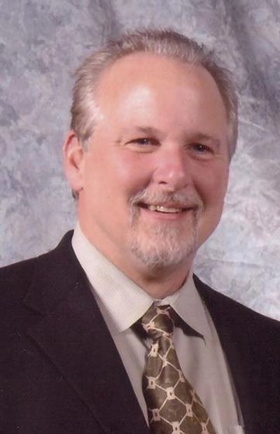 Smiling middle-aged man in a suit and patterned tie against a gray backdrop.