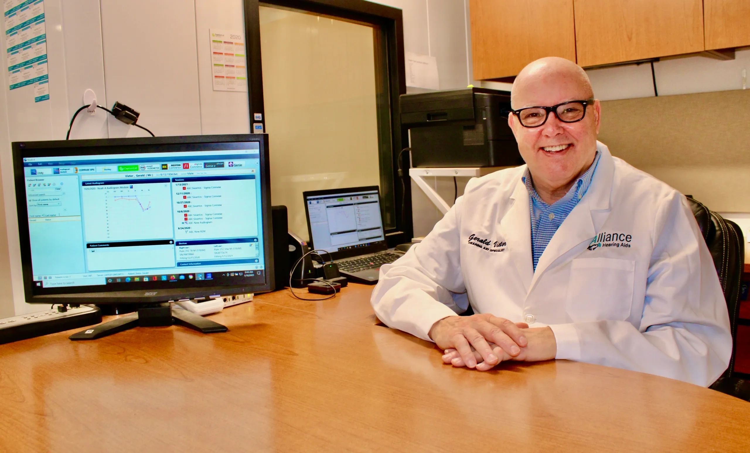 Smiling audiologist in a white coat at his desk with computer screens displaying hearing test data.