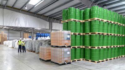 Workers inspect large stacks of green barrels and containers in a spacious warehouse.