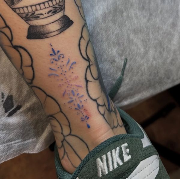 Delicate blue floral tattoo on ankle surrounded by larger black ink designs.