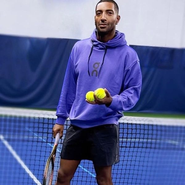 Man in purple hoodie holding two tennis balls on a tennis court.