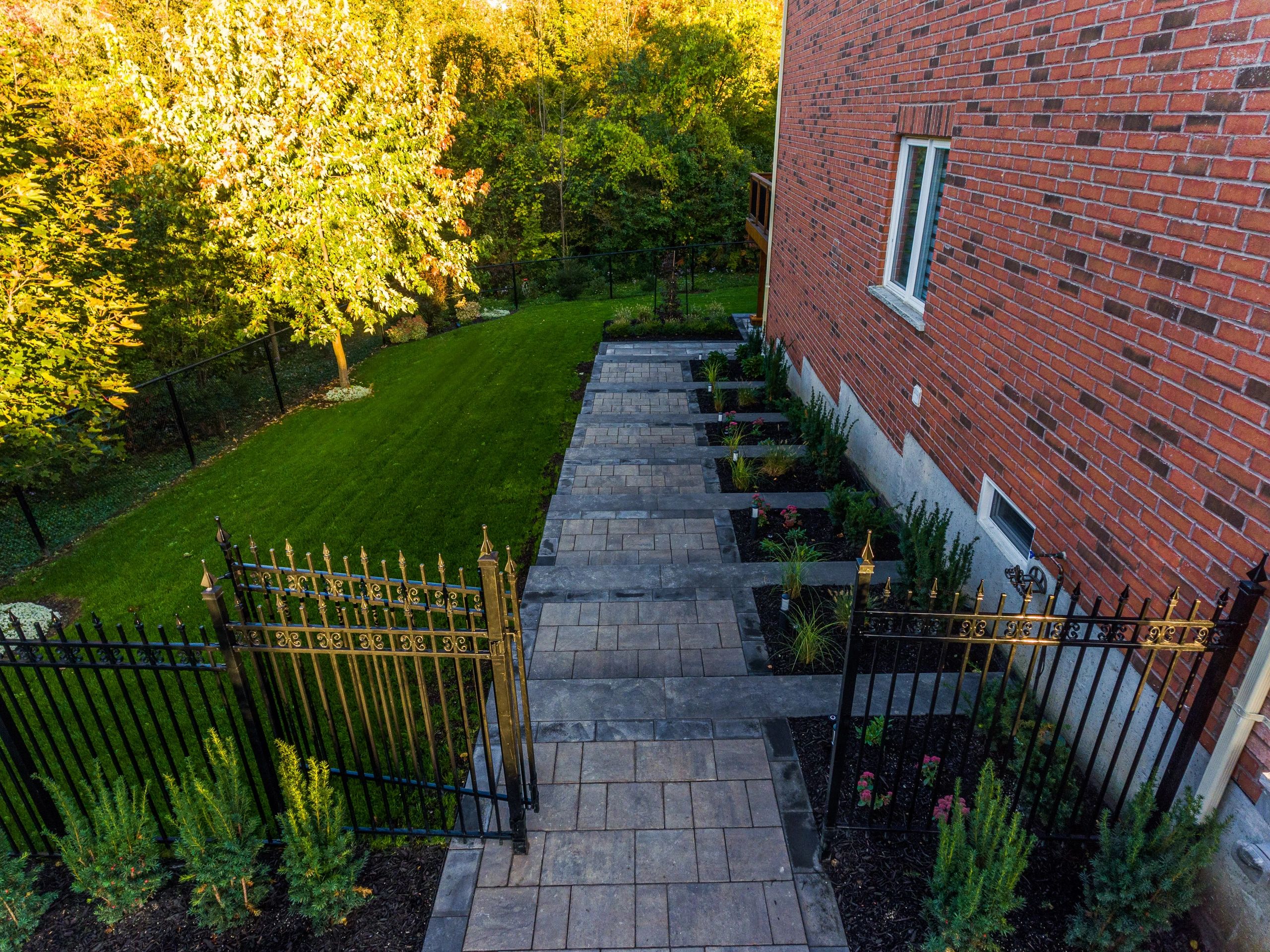 A paved walkway beside a brick house with garden beds and black iron gates.