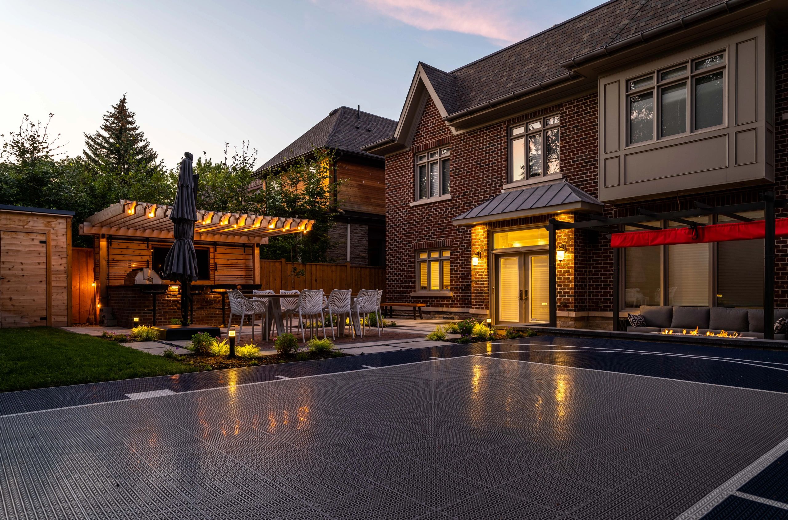 A cozy backyard with a basketball court, outdoor dining area, and brick house at dusk.