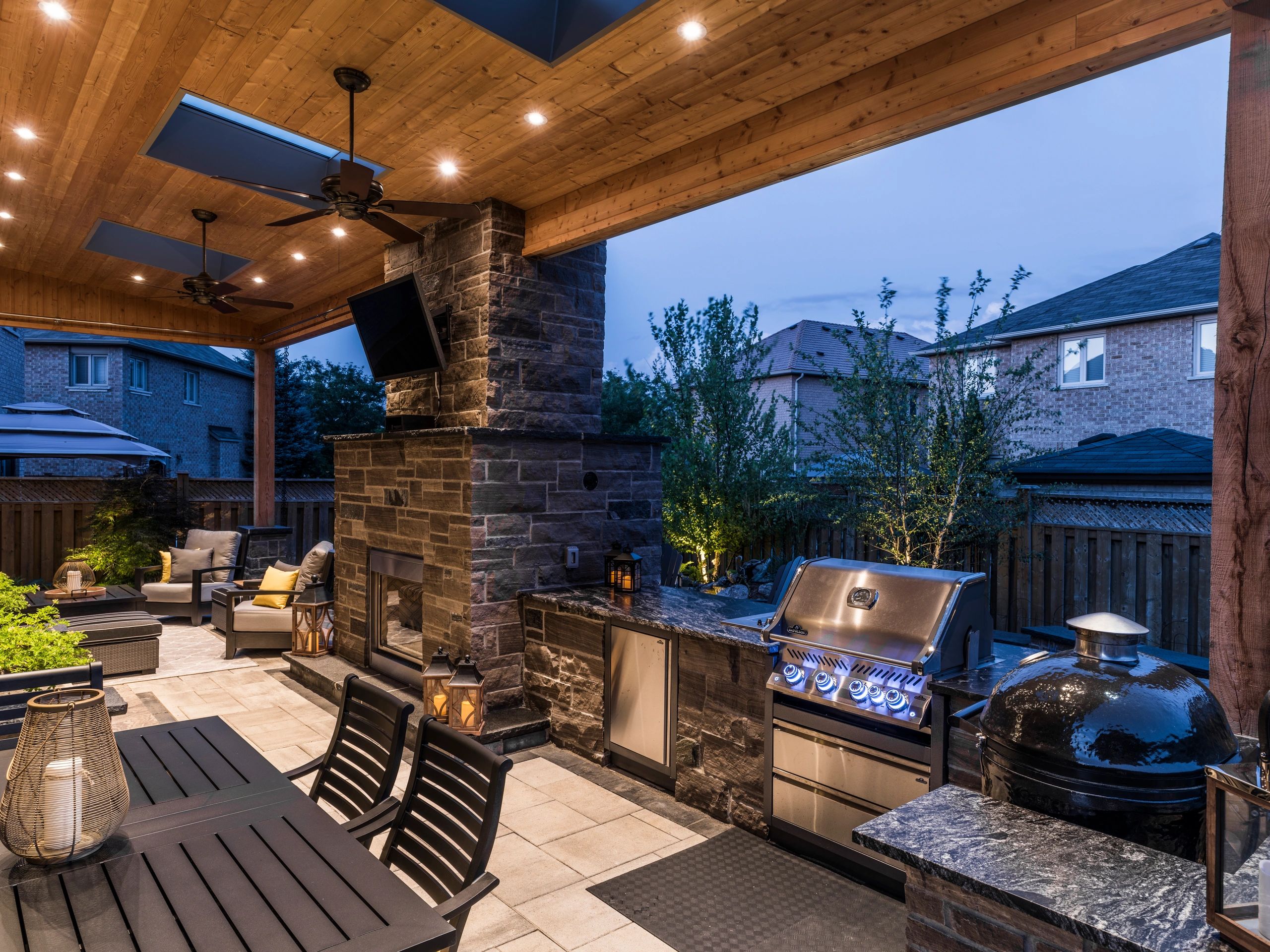 Cozy modern outdoor kitchen and seating area with stone fireplace and evening lighting.